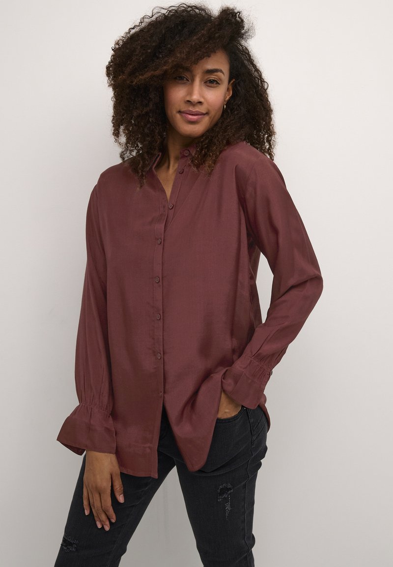 Camicia button-up bordeaux con una texture morbida e setosa, maniche lunghe con polsini arricciati e una vestibilità rilassata, indossata con jeans neri.