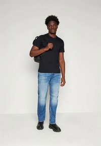 T-shirt en coton noir, jean denim bleu clair, bottes noires, modèle tenant un sac à dos noir, debout devant un fond blanc uni.