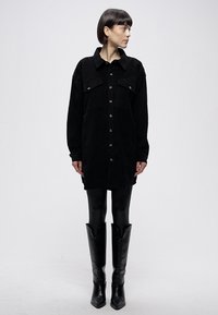 Svart manchester-skjortklänning med två bröstfickor, knappar framtill, långa ärmar och avslappnad passform, kombinerad med svarta högklackade stövlar.