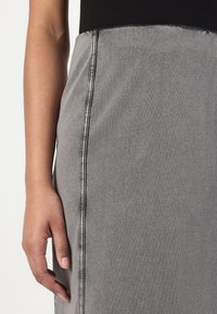 Fustă din denim gri, cu un textură fină, cu o croială dreaptă, având cusături verticale contrastante pe latură. Mână în prim-plan.