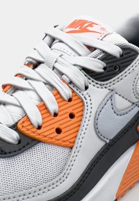 Nike tenisice s gornjim dijelom od mrežaste i gumene tkanine u bijeloj, narančastoj i sivoj boji, s teksturiranim narančastim detaljima i vezicama u ravnom tkanju.