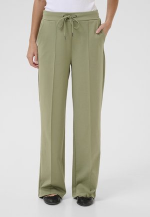 Vrouw die lichtgroene broek met brede pijpen draagt, met voorplooien, een taille met trekkoord en zijzakken, gecombineerd met zwarte ballerina's.