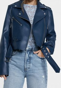 Veste de biker en simili cuir bleu marine avec des fermetures éclair argentées, des boutons à pression et des poignets ceinturés. Portée sur un haut en dentelle bleu clair et un jean en denim.