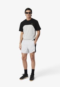 Zwart- en grijsgekleurde kleurblok T-shirt met korte mouwen, witte shorts, zwarte sneakers en een zonnebril. Bevat het Lacoste-logo op het shirt.
