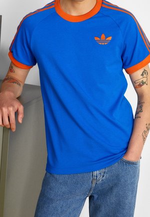 Osoba w niebieskiej koszulce Adidas z pomarańczowym obszyciem i paskami, niebieskie dżinsy oraz widoczne tatuaże na obu ramionach.