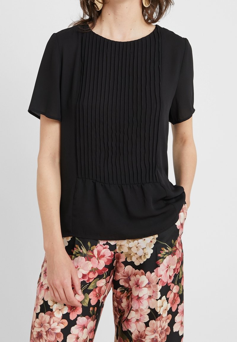Blouse noire à manches courtes avec des plis verticaux sur le devant et un ourlet froncé. Portée avec un pantalon à motif floral. Texture de tissu lisse.