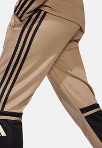 Beige Sporthosen mit schwarzen Seitenstreifen, ausgestattet mit einer Reißverschlusstasche und strukturierter Stoffqualität. Die Bündchen sind schwarz und sorgen für einen Farbkontrast.