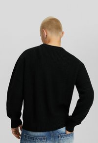 Osoba z krótkimi blond włosami, ubrana w czarny sweter z wełny i niebieskie dżinsy, pokazana od tyłu na jednolitym jasnym tle.