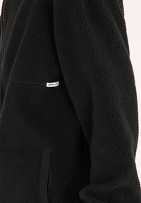 Pullover en polaire noir avec un tissu texturé, fermeture à moitié zippée et poche frontale. Il est doté d'une petite étiquette avec l'inscription "NEW BAD HABITS".