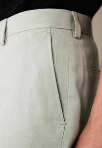 Light green trousers made of textured fabric with a smooth finish. Features a flat front, angled pockets, and a tailored waistband.