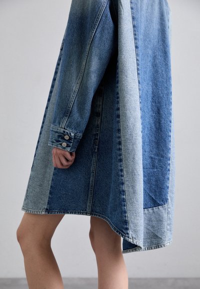 Džinsu mētelis, kas piedāvā tumši un gaiši zilā toņu kombināciju, ar brīvu piegriezumu, garām piedurknēm un kontrastējošiem audumiem sānos un apakšmalā.