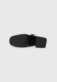 Semelle de chaussure noire avec un motif de grip texturé, de forme rectangulaire et un talon légèrement surélevé. Le matériau semble être en caoutchouc avec des bords lisses.