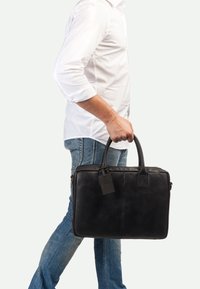Schwarze Lederaktentasche mit doppelten Griffen, rechteckiger Form, glatter Oberfläche, Etikett und Reißverschluss, gehalten von einer Person in Jeans und einem weißen Hemd.