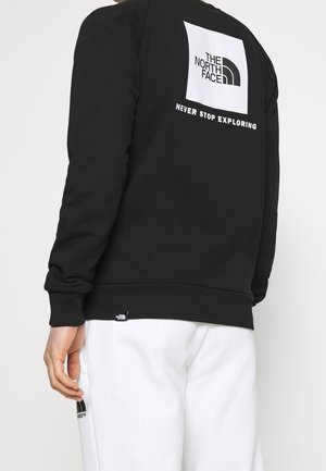 Person trägt schwarzen Sweatshirt mit weißem "The North Face"-Logo und dem Text "Never Stop Exploring" auf dem Rücken, kombiniert mit weißer Hose.