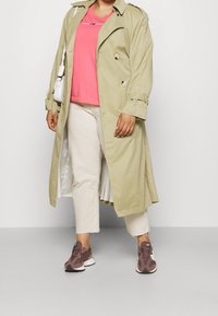 Trench coat verde claro, camiseta rosa, bolsa blanca, pantalones cortos blancos y zapatillas marrones con malla texturizada y acentos sólidos.