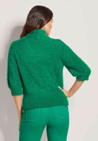 Grüner, flauschiger Pullover mit hohem Kragen und Dreiviertelärmeln, kombiniert mit passenden grünen Hosen, die eine weiche Textur und eine lockere Passform zeigen.