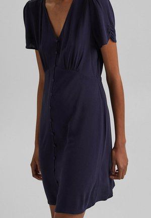 Kobieta w granatowej sukience z krótkimi rękawami, z przodu zapinanej na guziki i z marszczeniami na rękawach, na jednolitym tle.