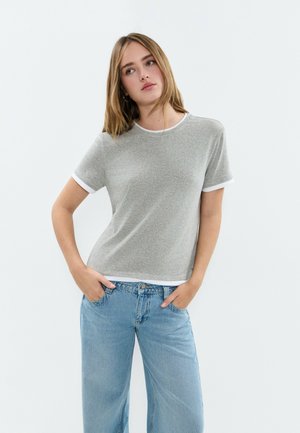 Joven con cabello lacio que lleva una camiseta gris de manga corta con ribete blanco y pantalones anchos azul claro, con las manos en los bolsillos, fondo neutro.