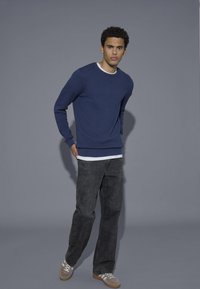 Navy textured sweater with ribbed cuffs and hem, layered over a white shirt, paired with dark gray jeans and beige sneakers.