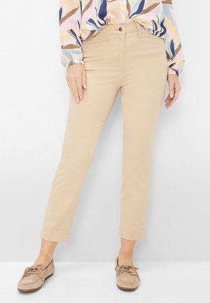 Frau trägt beigefarbene, gekürzte Hose, gemustertes Langarmhemd, in die Hose gesteckt, sowie passende beige Schnür-Loafers, steht vor einer weißen Wand.