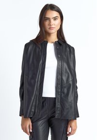 Sort læderjakke med krave, knapper foran og paneldetaljer. Glat tekstur; båret over en hvid skjorte.