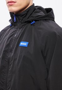 Jachetă ușoară neagră cu glugă, având un logo albastru de marcă, fermoar contrastant și șnururi reglabile la nivelul gâtului.