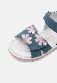 Sándalo de ante azul con diseño de flores de purpurina rosa, correa de velcro y suela blanca con un patrón de estrellas rosas y plateadas en la plantilla.
