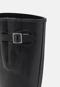 Botte en caoutchouc noire présentant une texture lisse, un haut court et une accent en boucle argentée sur le côté. L'intérieur est doublé d'un tissu doux.