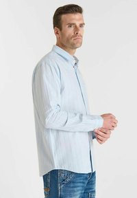 Man in een lichtblauw gestreept overhemd met knopen en blauwe jeans die zijn manchetten aanpast, terwijl hij naar links kijkt tegen een eenvoudige witte achtergrond.
