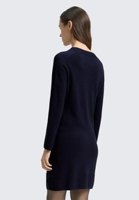 Granatowa dzianinowa sukienka z długimi rękawami, prążkowanymi detalami przy mankietach i brzegu oraz odkrytymi plecami. Gładka faktura, dopasowana sylwetka.