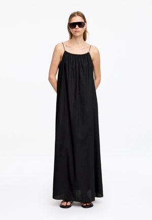 Femme portant une robe maxi noire ample sans manches, de grandes lunettes de soleil foncées et des sandales noires, debout les mains dans le dos.
