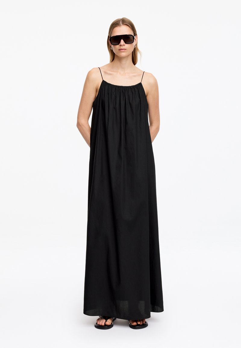 Femme portant une robe maxi noire ample sans manches, de grandes lunettes de soleil foncées et des sandales noires, debout les mains dans le dos.