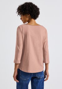 Top rosa claro de manga tres cuartos con escote en la espalda en forma de barco, hecho de una tela suave, combinado con jeans de mezclilla azules.