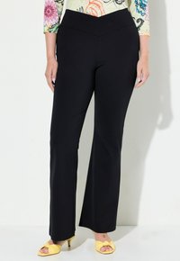 Pantalons noirs ajustés avec une taille large, présentant une texture lisse et des jambes évasées. Associés à des sandales jaunes nouées.