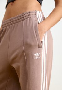Adidas trenirke v bežno-rjavi barvi, ki imajo elastičen pas, stranske žepke in bele tri stripe z logotipom.