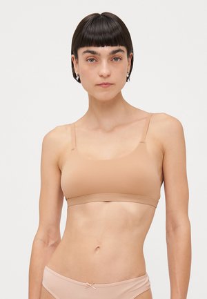 Femme aux cheveux courts et foncés portant un soutien-gorge et des sous-vêtements couleur chair, sur fond clair uni, regardant droit devant avec une expression neutre.