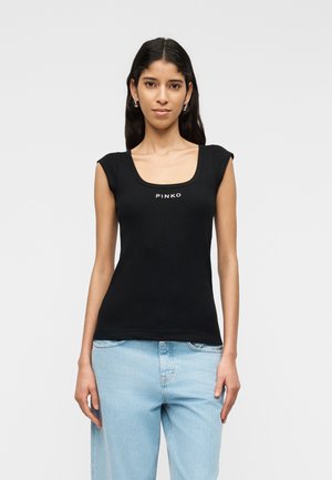 Giovane donna che indossa un top nero senza maniche con la scritta "PINKO" e jeans azzurri, in piedi davanti a uno sfondo semplice di colore grigio chiaro.