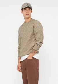 Maglione lavorato a maglia marrone, con scollo tondo, indossato sopra una camicia bianca, abbinato a pantaloni marroni e un cappellino grigio chiaro.