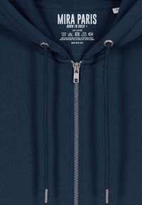 Felpa blu in 100% cotone con zip frontale, cordini e un'etichetta che mostra "MIRA PARIS", istruzioni per la cura e un logo.