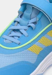 Detalhe de um tênis azul com cadarços azul-claro, riscas amarelas, acabamentos verdes e sola branca.