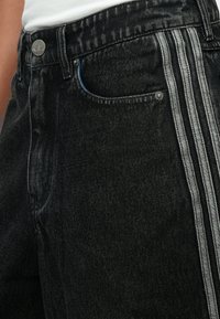 Μαύρο τζιν με ελαφρώς ξεθωριασμένη υφή, με τρεις λευκές πλευρικές ρίγες και ένα μεταλλικό κουμπί με λογότυπο στη μέση.