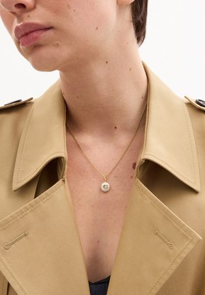 Primo piano di una persona che indossa un trench beige e una collana d'oro con un ciondolo bianco che presenta una lettera "C" dorata.