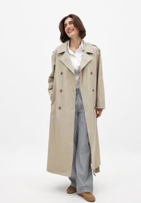 Bēša trenažieris ar diviem pogām priekšpusē, platām atlokām un plecu epauletēm. Apvienots ar svītrainām platām biksēm un brūniem apaviem.