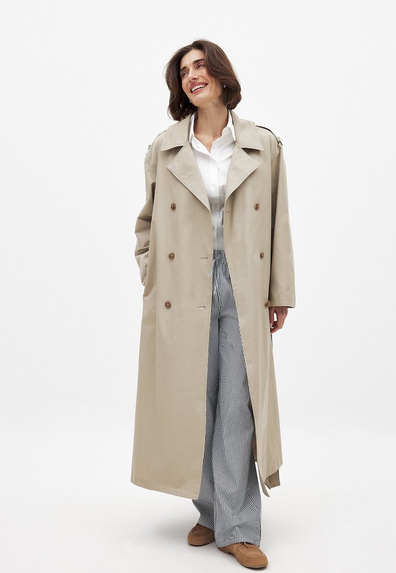 Bēša trenažieris ar diviem pogām priekšpusē, platām atlokām un plecu epauletēm. Apvienots ar svītrainām platām biksēm un brūniem apaviem.