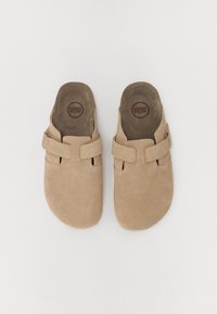 Sabots en daim beige avec une texture lisse, une forme arrondie et deux sangles réglables sur le dessus pour un ajustement sécurisé.