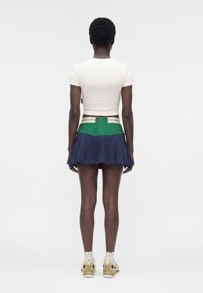 Γυναίκα με πλάτη φοράει λευκό crop top, πράσινη και μπλε πλισέ μίνι φούστα και μπεζ αθλητικά παπούτσια σε απλό λευκό φόντο.
