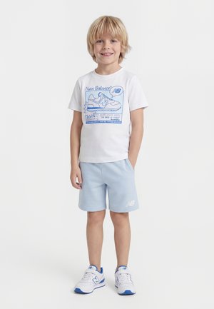 Niño con cabello rubio que lleva una camiseta blanca de New Balance, pantalones cortos azul claro y zapatillas blancas con detalles en azul, de pie y sonriendo.