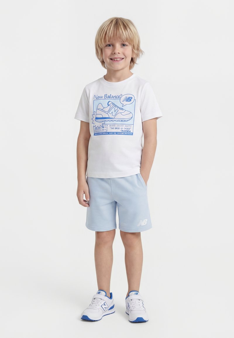 Αγόρι με ξανθά μαλλιά που φοράει ένα λευκό t-shirt New Balance, ανοιχτό μπλε σορτς και λευκά αθλητικά παπούτσια με μπλε λεπτομέρειες, στέκεται και χαμογελά.