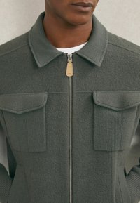 Jachetă tricotată verde cu fermoar frontal, două buzunare pe piept și manșete elastice. Are un guler structurat și un mâner de fermoar din piele.