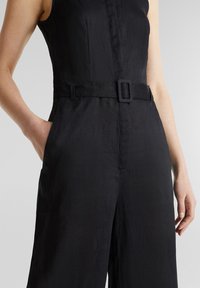 Combinaison noire sans manches avec ceinture en tissu et boucle, les mains reposant sur les côtés, sur un fond gris neutre.